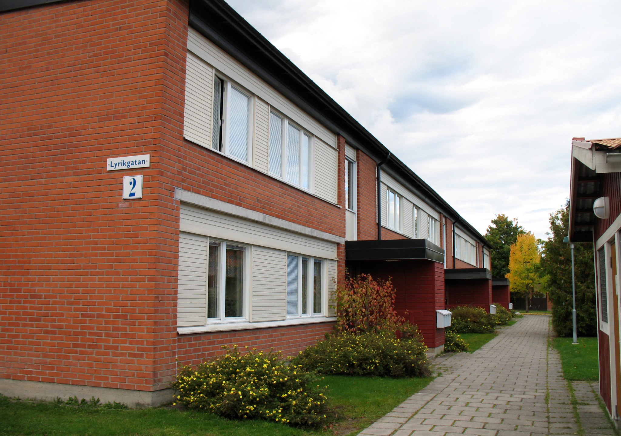 Tegel lägenhetsbyggnad med vita fönsterluckor, skylt "Lyrikgatan 2," och trädgårdsväg.