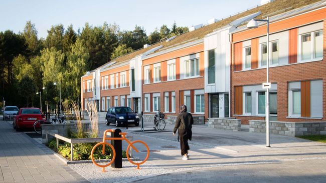 Modern bostadsområde med röda tegelbyggnader, parkerade bilar och en person som går på trottoaren.