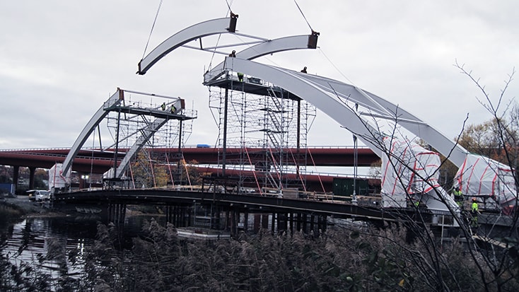 Byggnadsarbetare monterar en modern bågbro över en flod mitt bland byggnadsställningar och kranar.