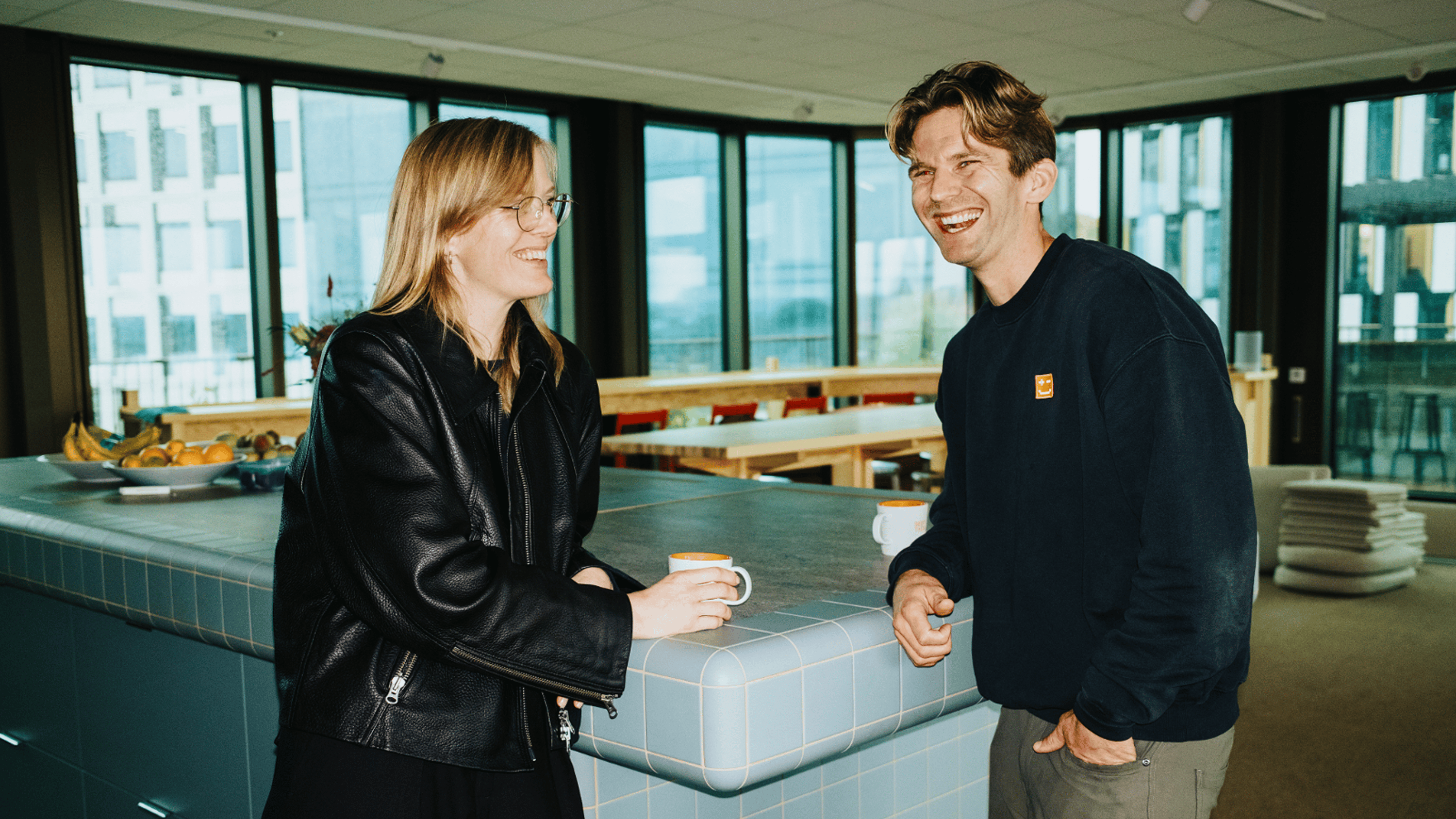 Två personer som dricker kaffe och samtalar i ett modernt kontorskök med stora fönster.