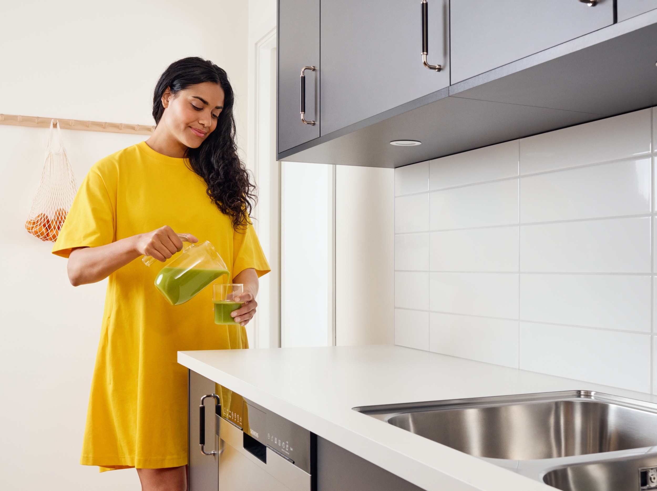 Kvinna i gul klänning häller grön juice i modernt kök med grå skåp och vita kakel.