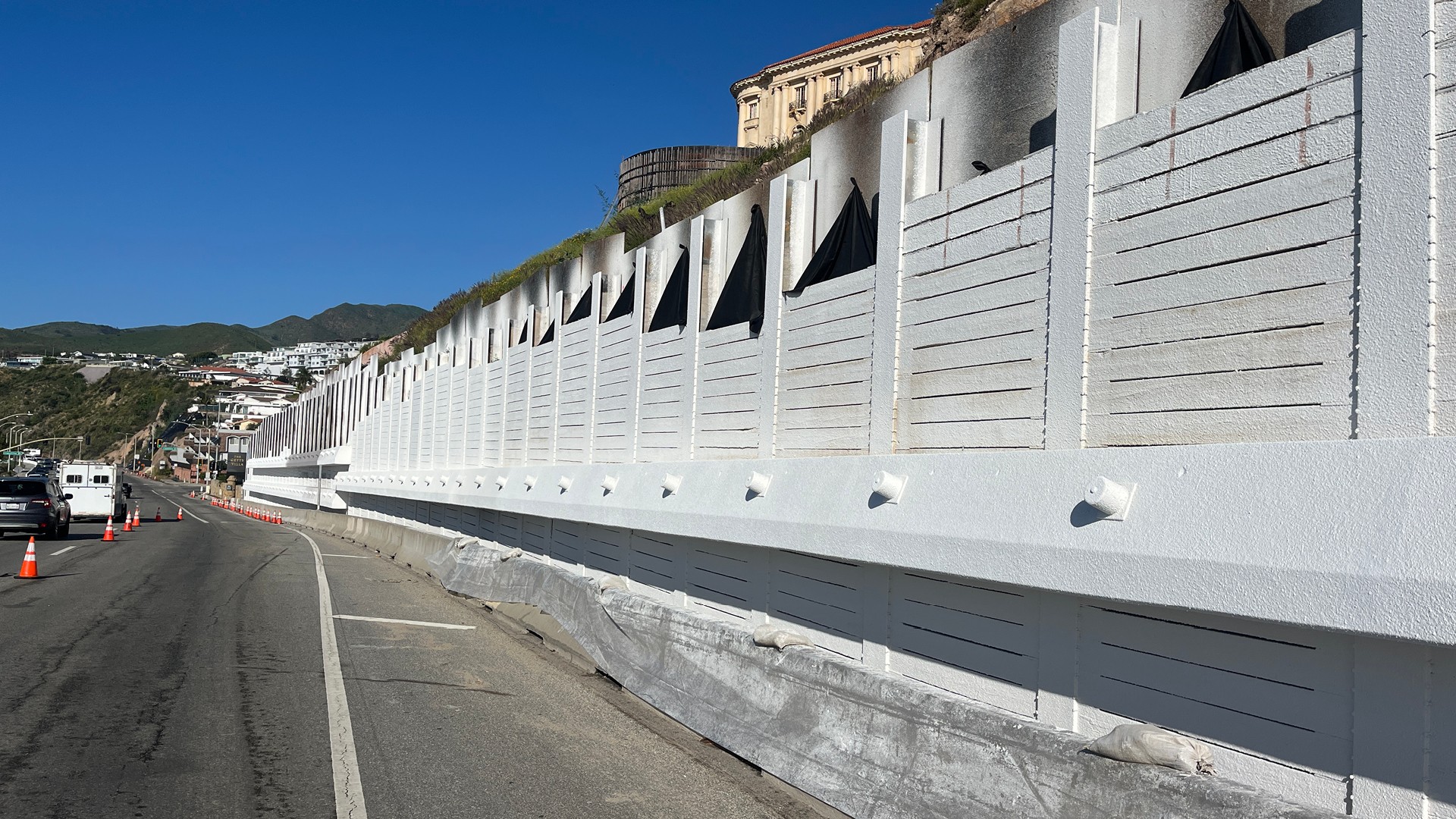 Fire-Resistant Retaining Wall Repairs Near Getty Villa Long white sound barrier wall along a coastal road with hills and buildings in the background.