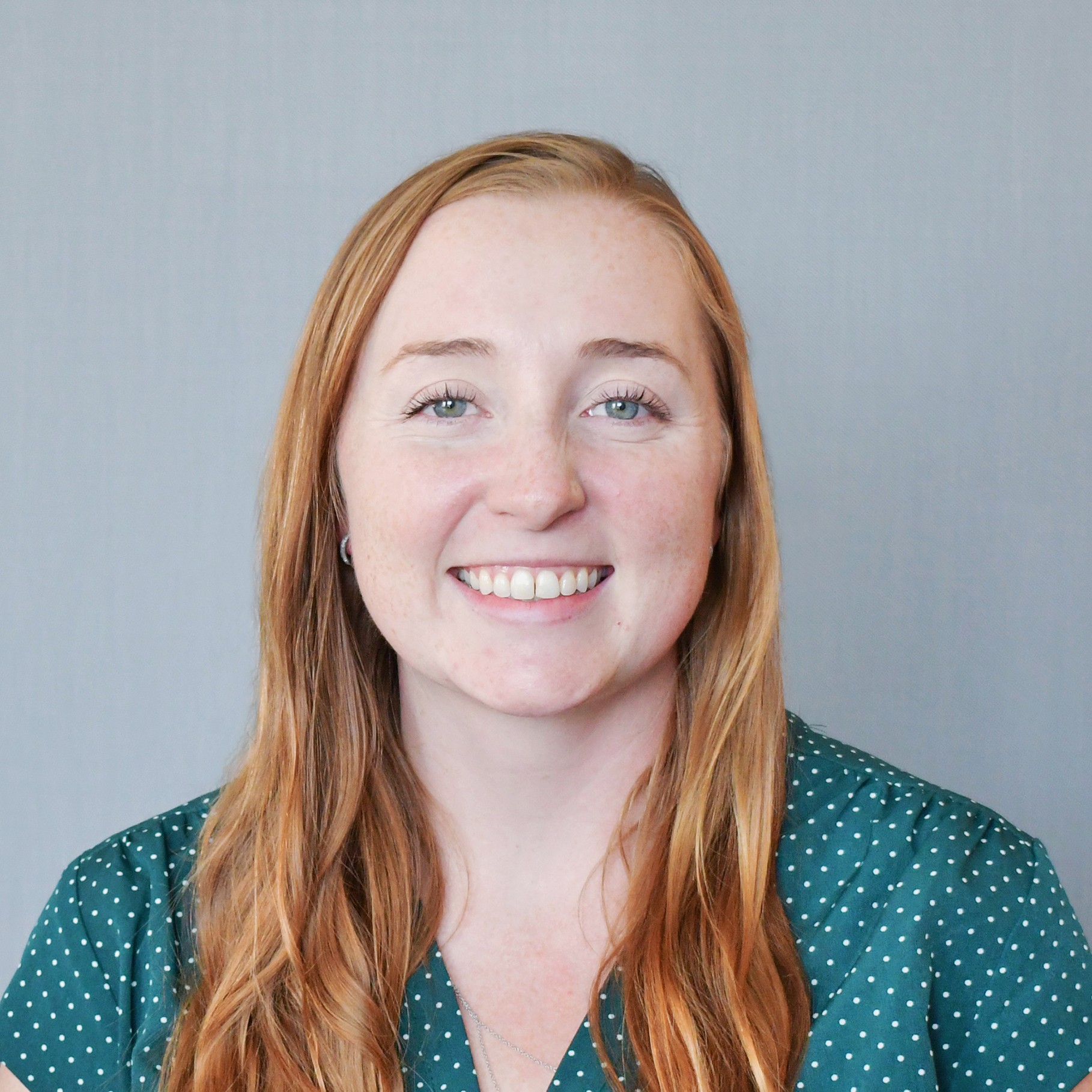 Woman with long red hair wearing a green polka dot blouse against a gray background.