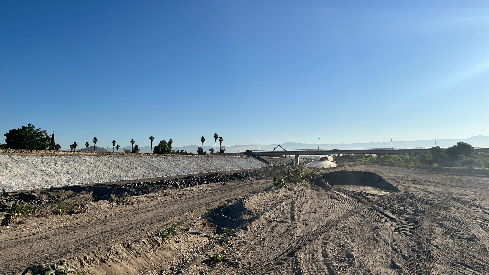 Riverside Levees Rehabilitation Along the Santa Ana River Dry riverbed with dirt tracks and a concrete-lined section under a clear blue sky.