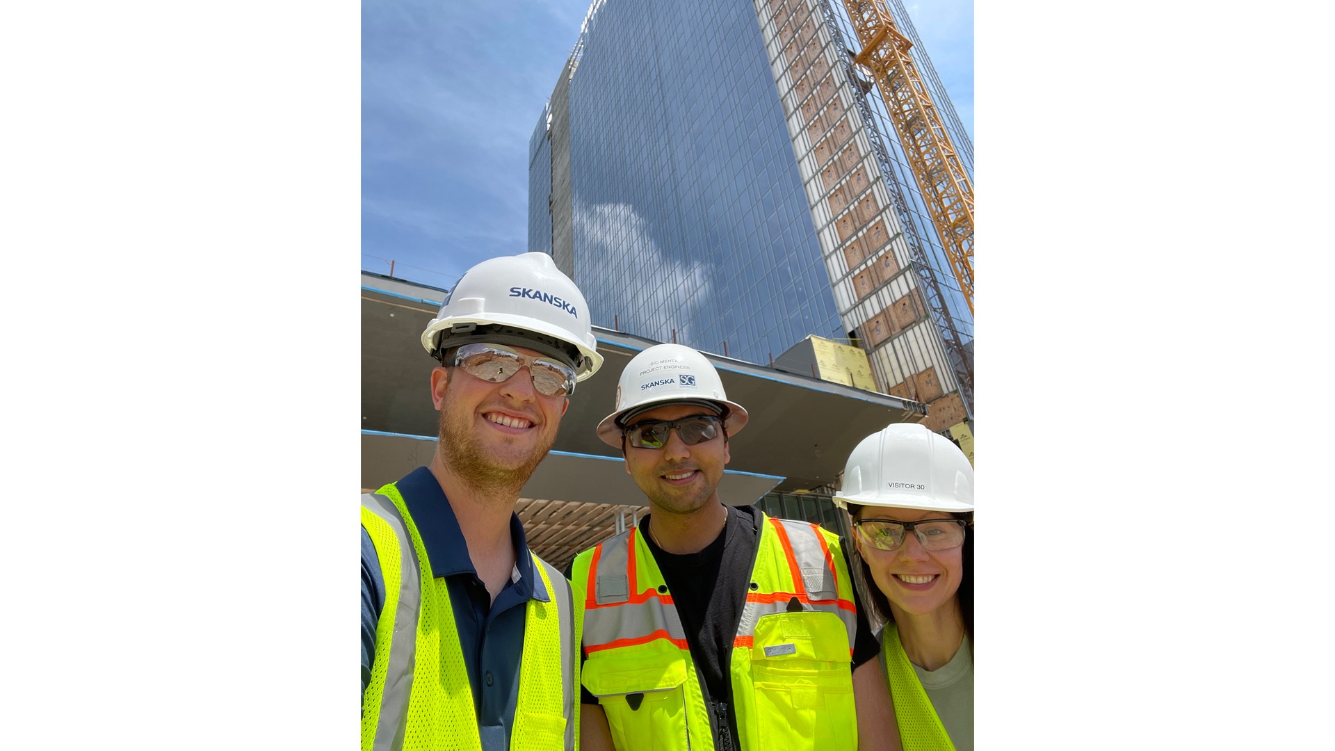 Construction workers in safety gear at urban high-rise site Three construction workers wearing safety vests and helmets stand in front of a high-rise building.