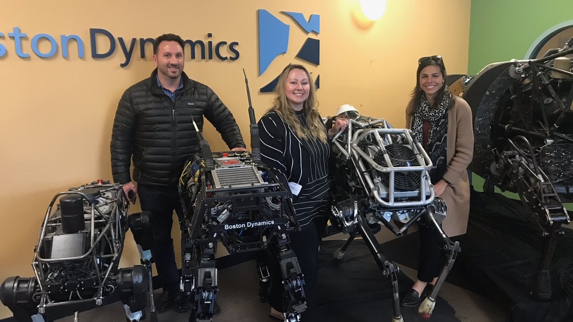 Boston Dynamics Robots with Team Members in Tech Showcase Three people stand with Boston Dynamics robotic quadrupeds in a tech exhibit room.
