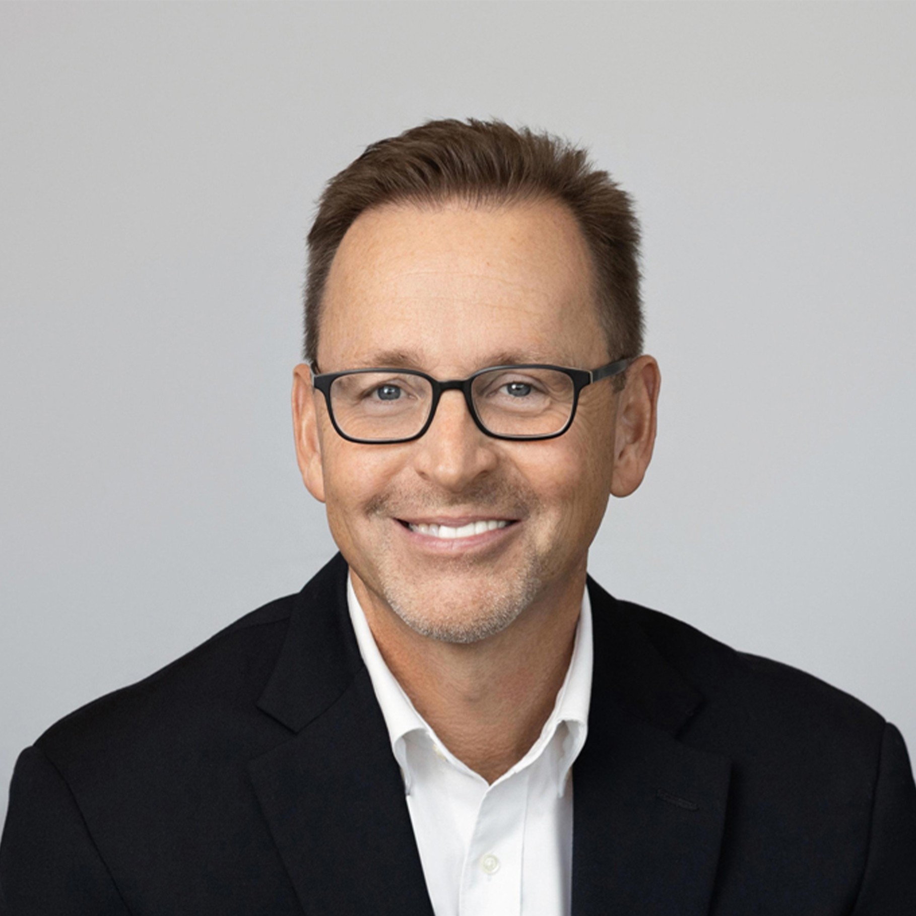 A man in a black suit jacket and white shirt sits against a plain gray background.