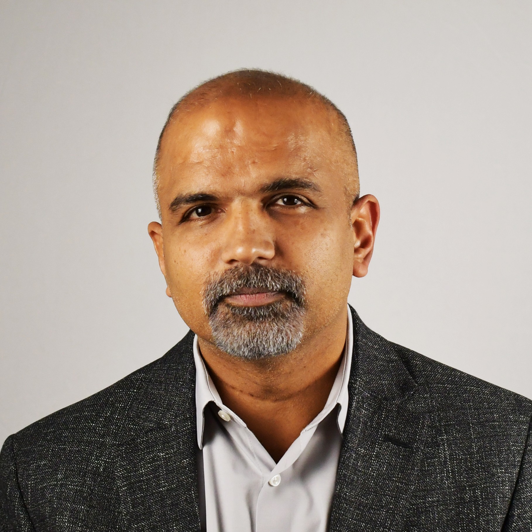 A man in a gray textured suit jacket and white shirt against a neutral background.