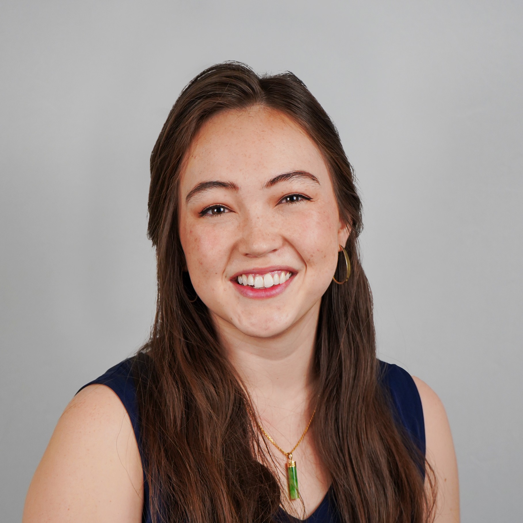 Woman with long brown hair wearing a navy blue top and green pendant necklace.