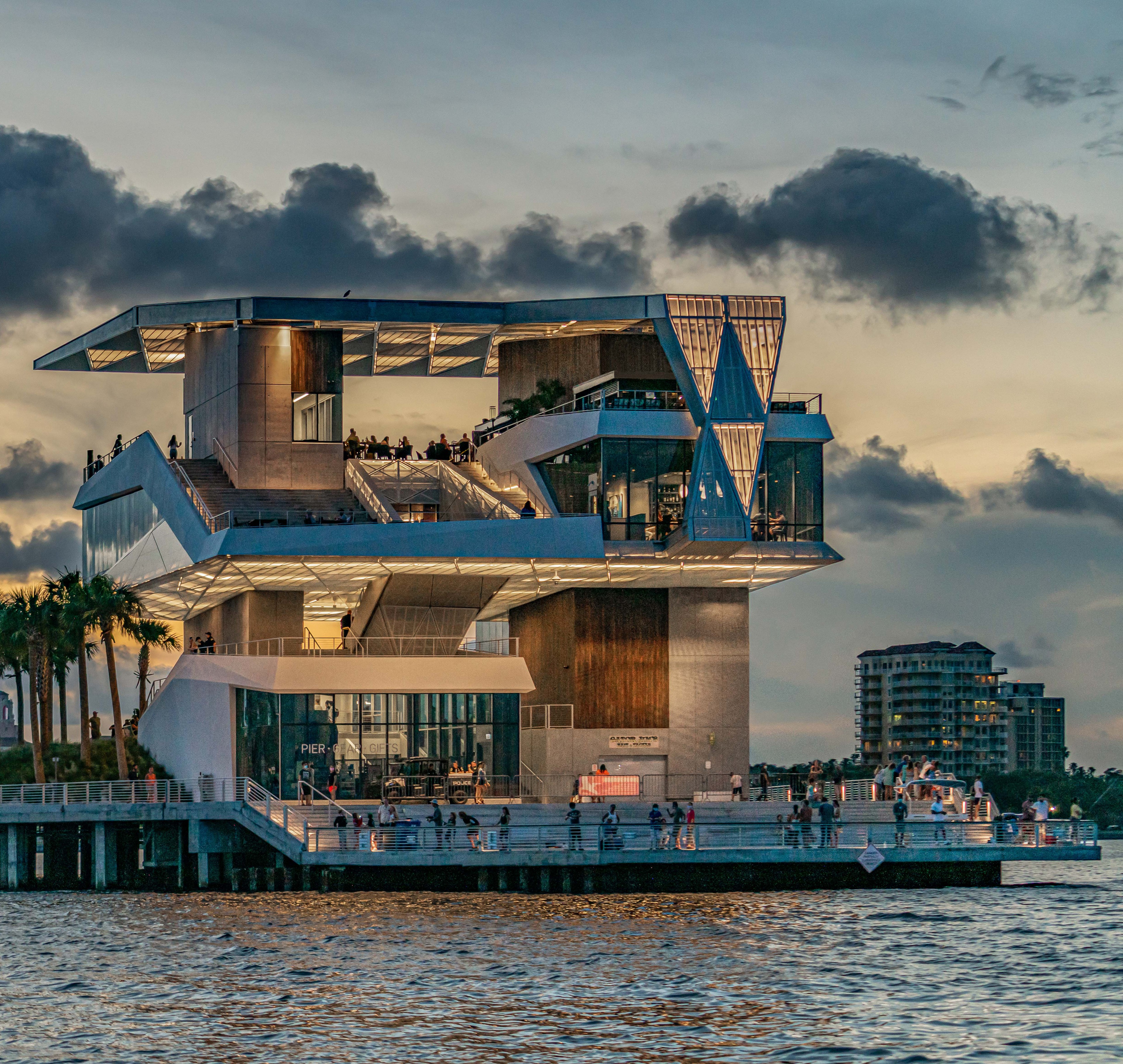 Modern waterfront building with palm trees and people at sunset over calm water.