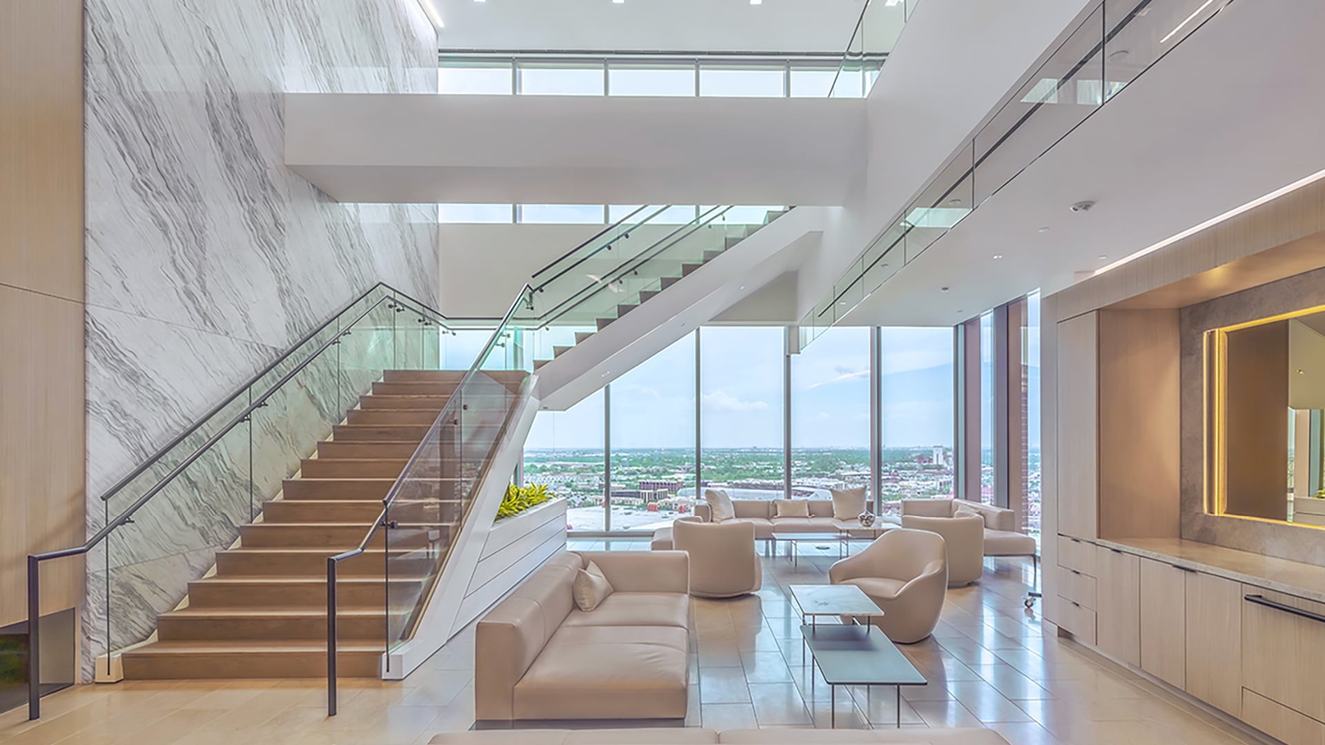 Modern office lounge with glass staircase, beige seating, and large city-view windows.