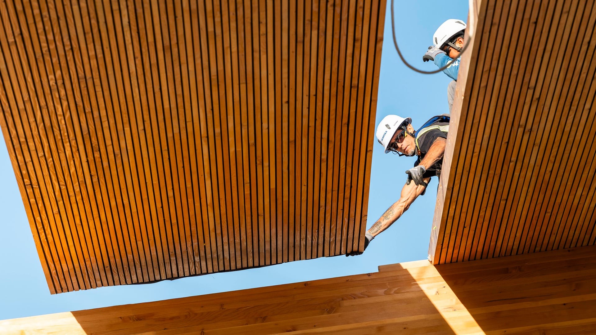 Two construction workers in helmets installing large wooden panels under clear blue sky.