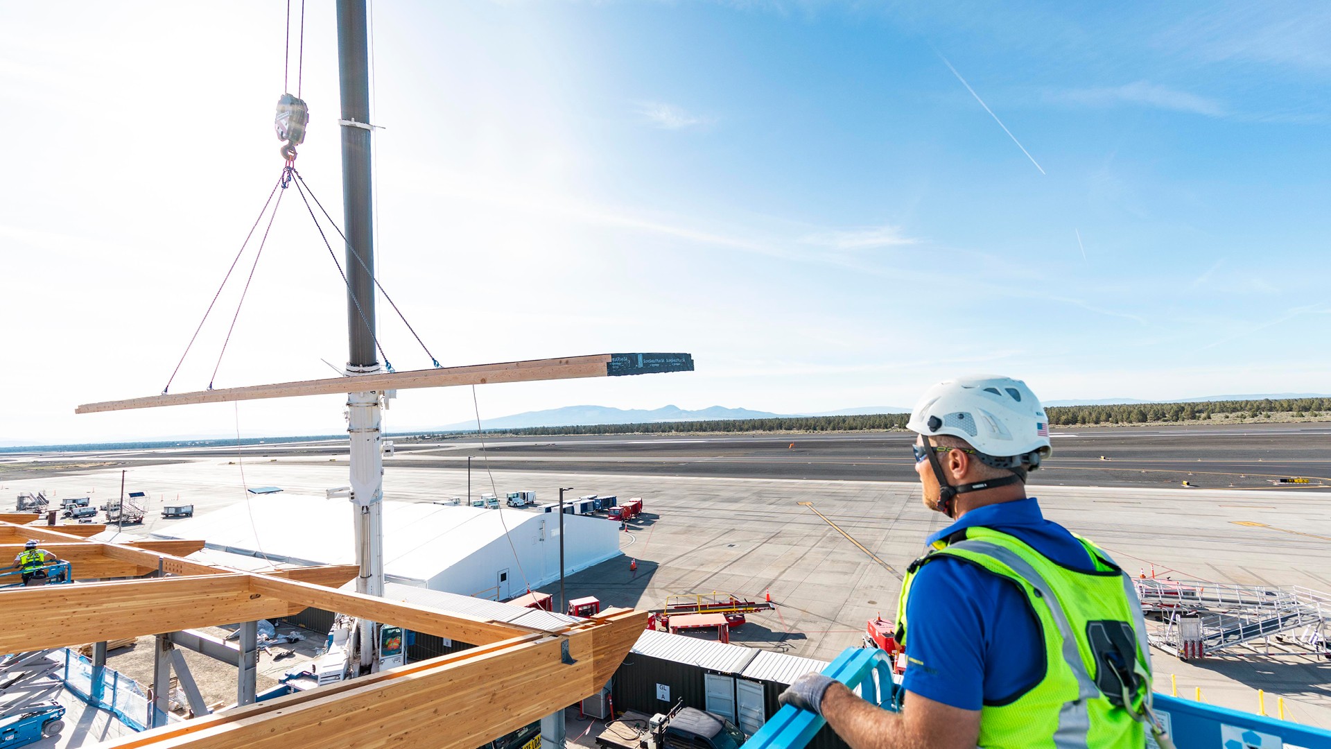 Craftworkers Position Mass Timber Roof Panels During Installation Construction worker in safety gear overseeing wooden beam installation at airport site.