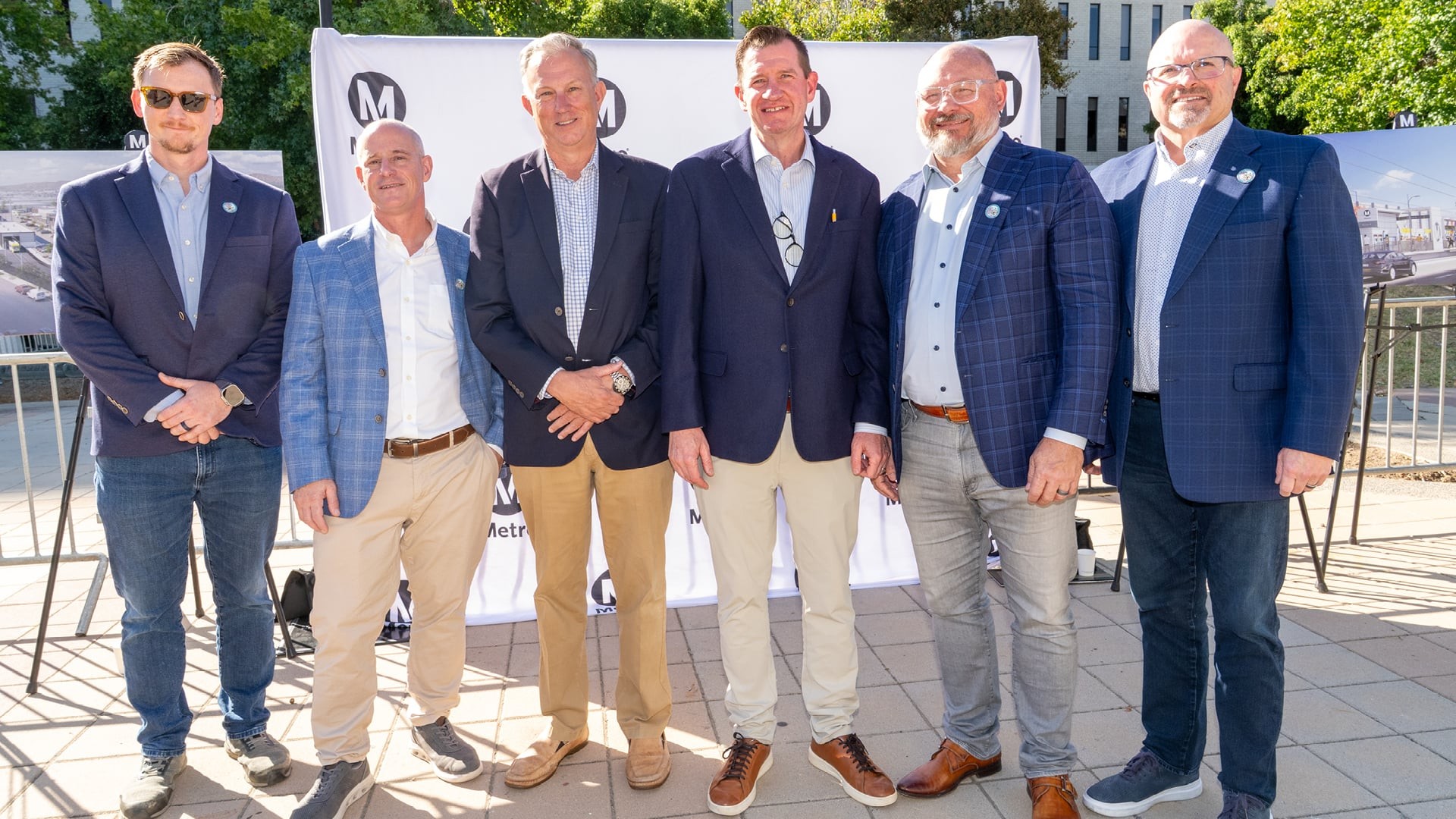 East San Fernando Valley Light Rail Transit Project Groundbreaking Six men in business casual attire pose outdoors at a Metro event with banners behind.