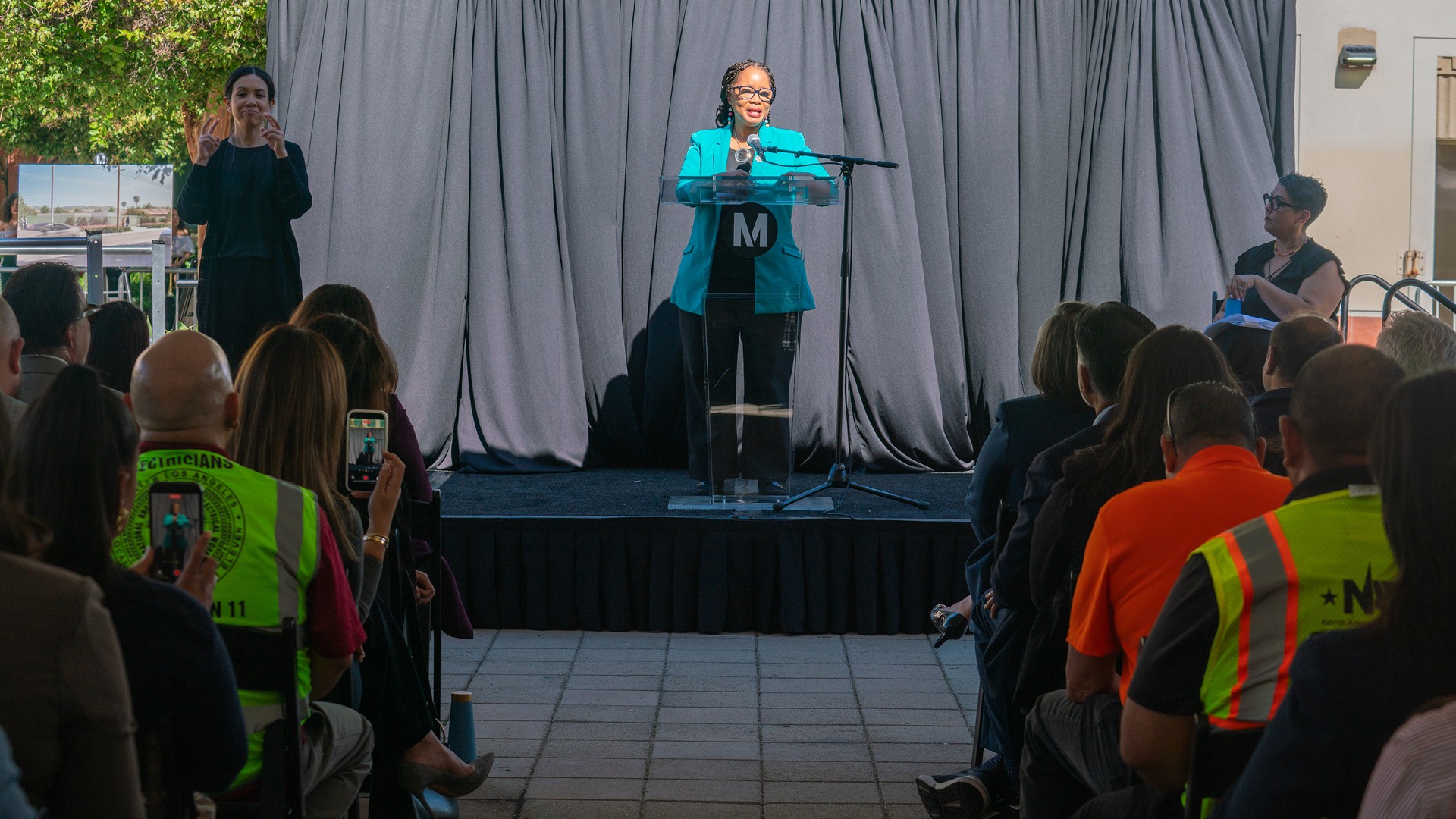 East San Fernando Valley Light Rail Transit Project Groundbreaking Woman in teal jacket speaking at a podium during an indoor event with audience.