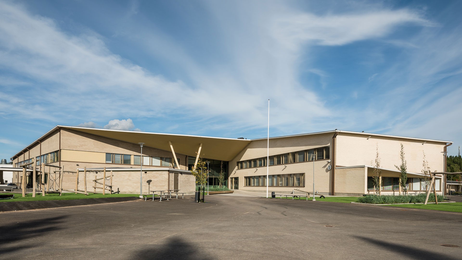 Pihalla on turvalliset leikki- ja oleskelupihat. Moderni koulurakennus kulmikkaalla katolla ja suurilla ikkunoilla, taustanaan kirkas sininen taivas.