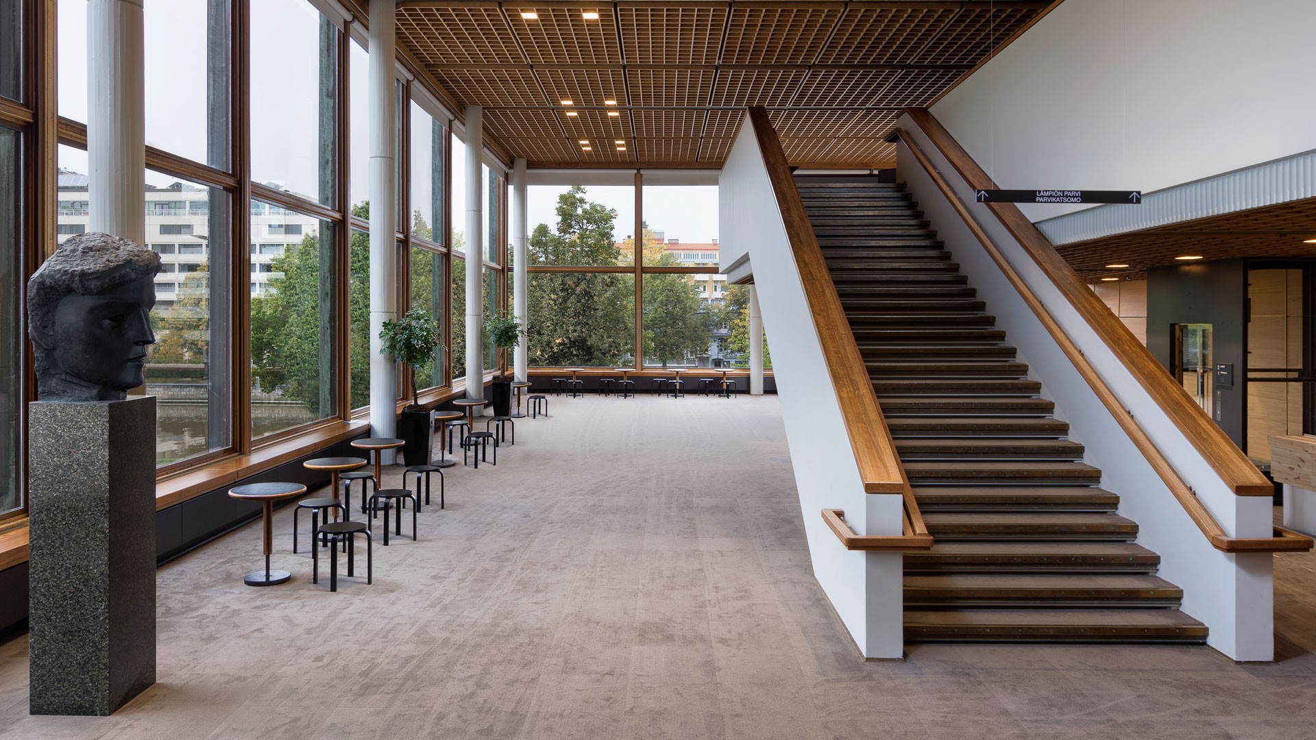 Moderni aula suurilla ikkunoilla, puiset portaat, istuinalue ja veistos jalustalla.