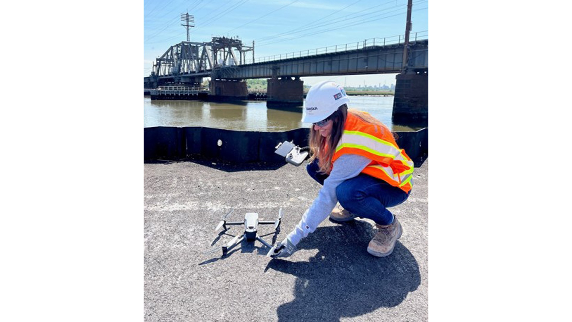 Person in safety gear operating a drone near a river with a bridge in the background.