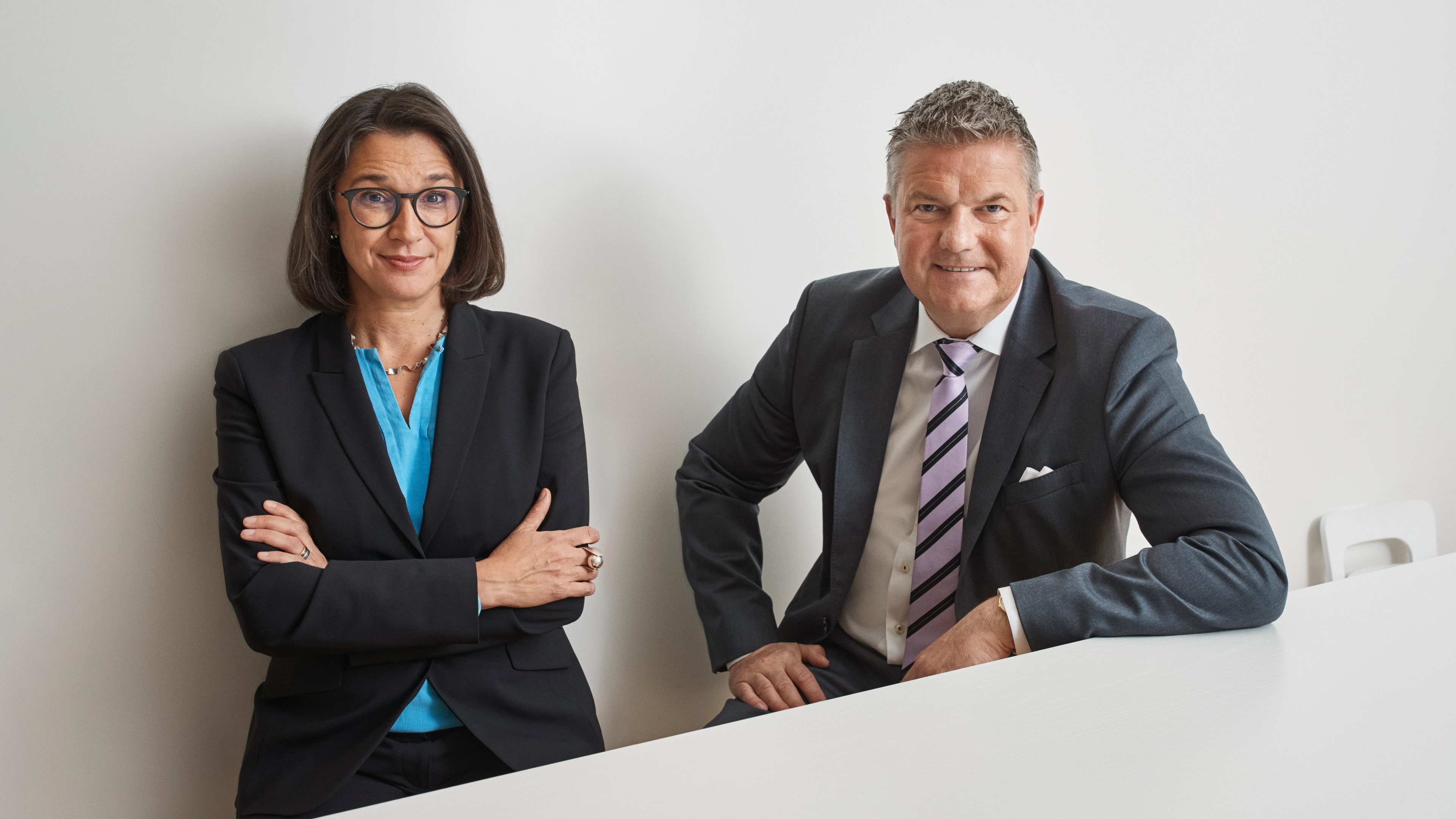 Lena Hök and Anders Danielsson Two business professionals in formal suits sitting at a white table against a plain wall.