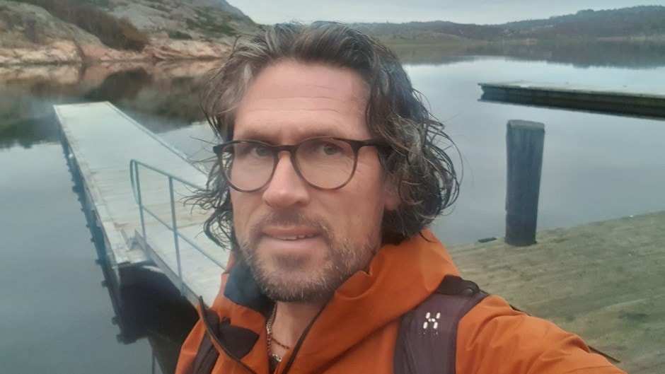 Person i en orange jacka som tar en selfie på en brygga vid lugnt sjövatten med kullar.