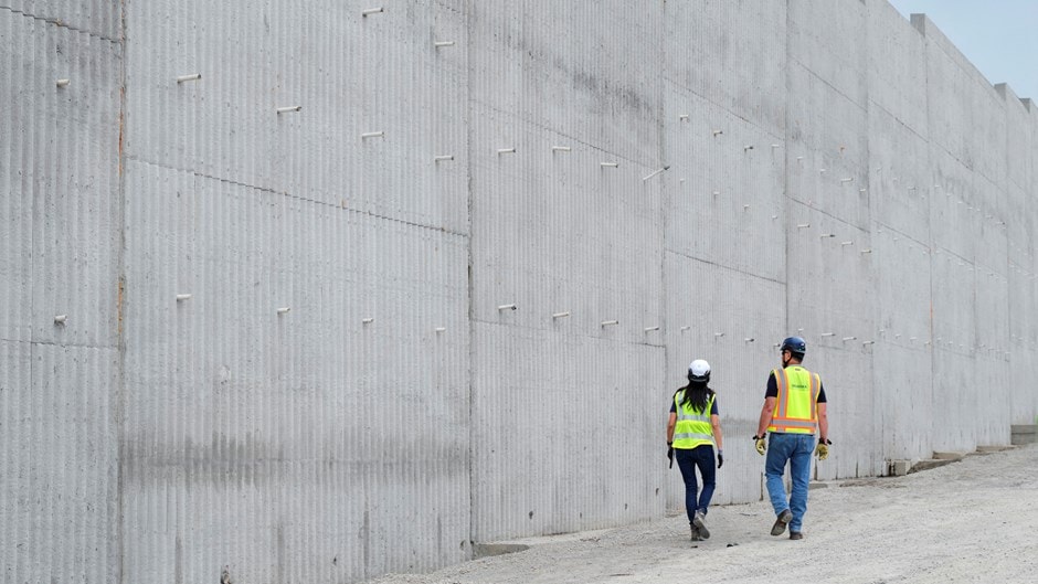 Två byggnadsarbetare i säkerhetsutrustning går längs en stor betongvägg på en arbetsplats.
