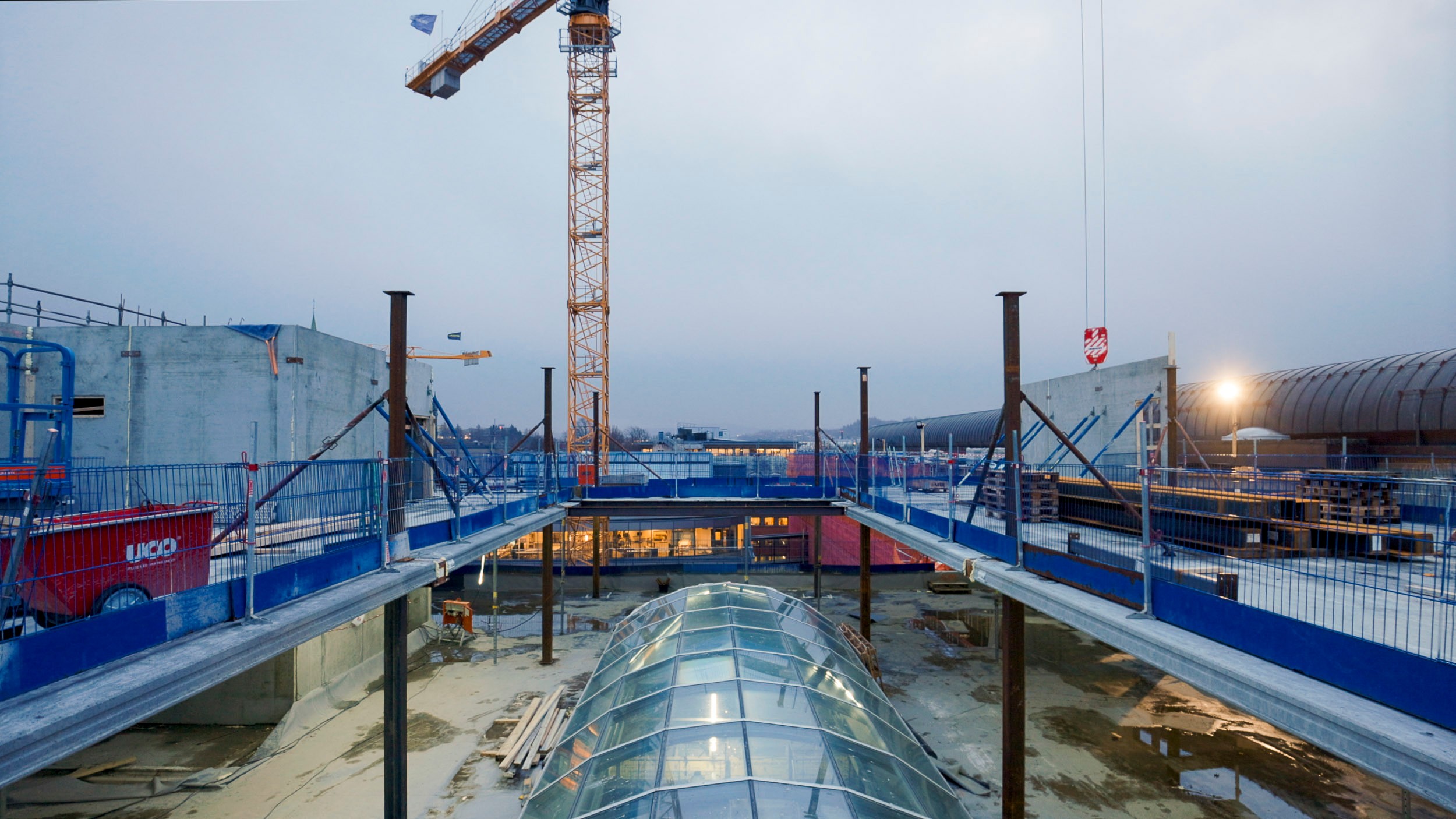 Byggeplass med stålkonstruksjon, kran og glass-tak under overskyet himmel.