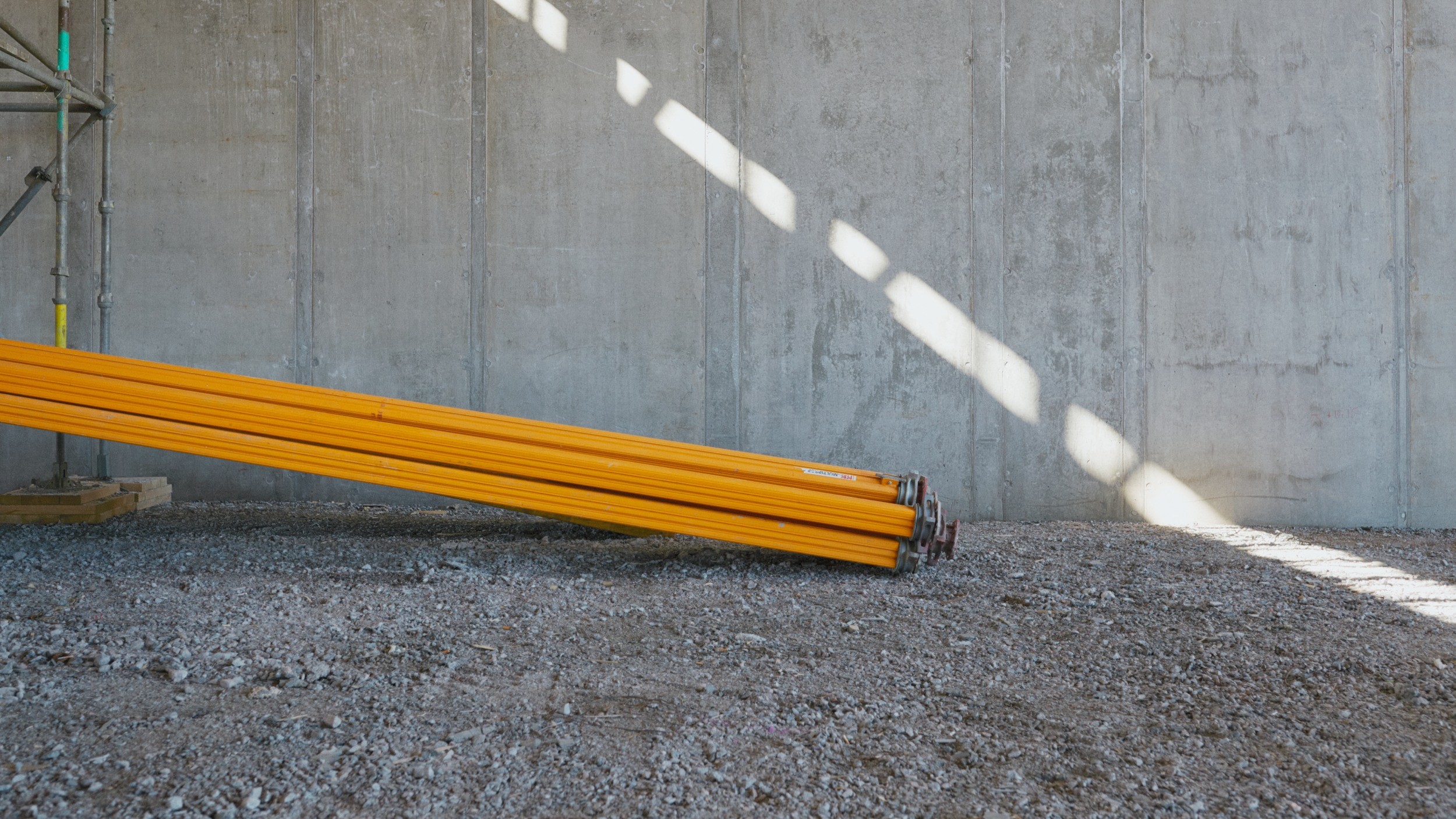 Yellow metal pipes stacked on gravel, leaning against a concrete wall at a construction site.