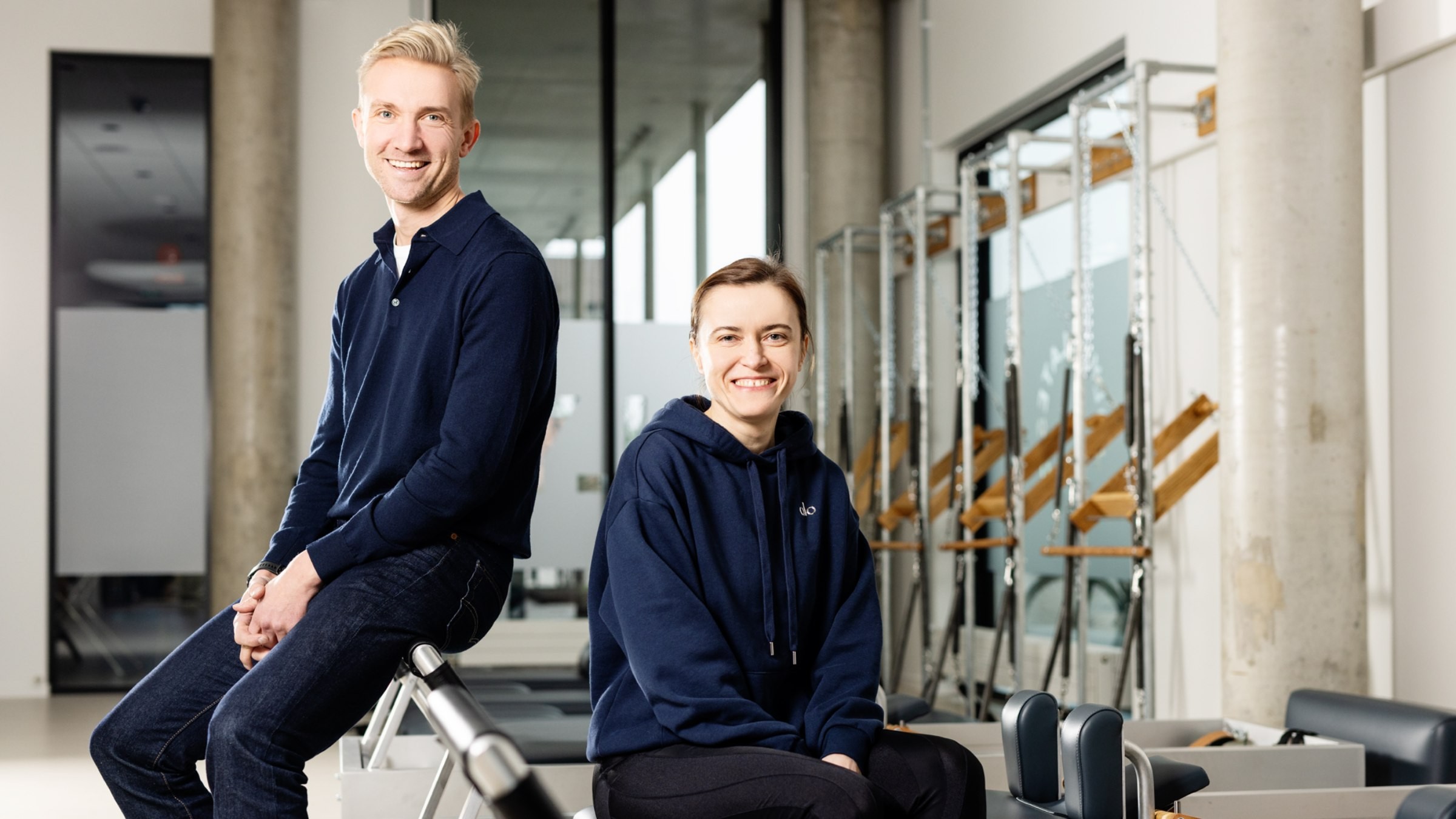 Modern fitnessstudio med pilatesutrustning Två personer i avslappnad klädsel sitter i ett modernt gym med träningsutrustning i bakgrunden.