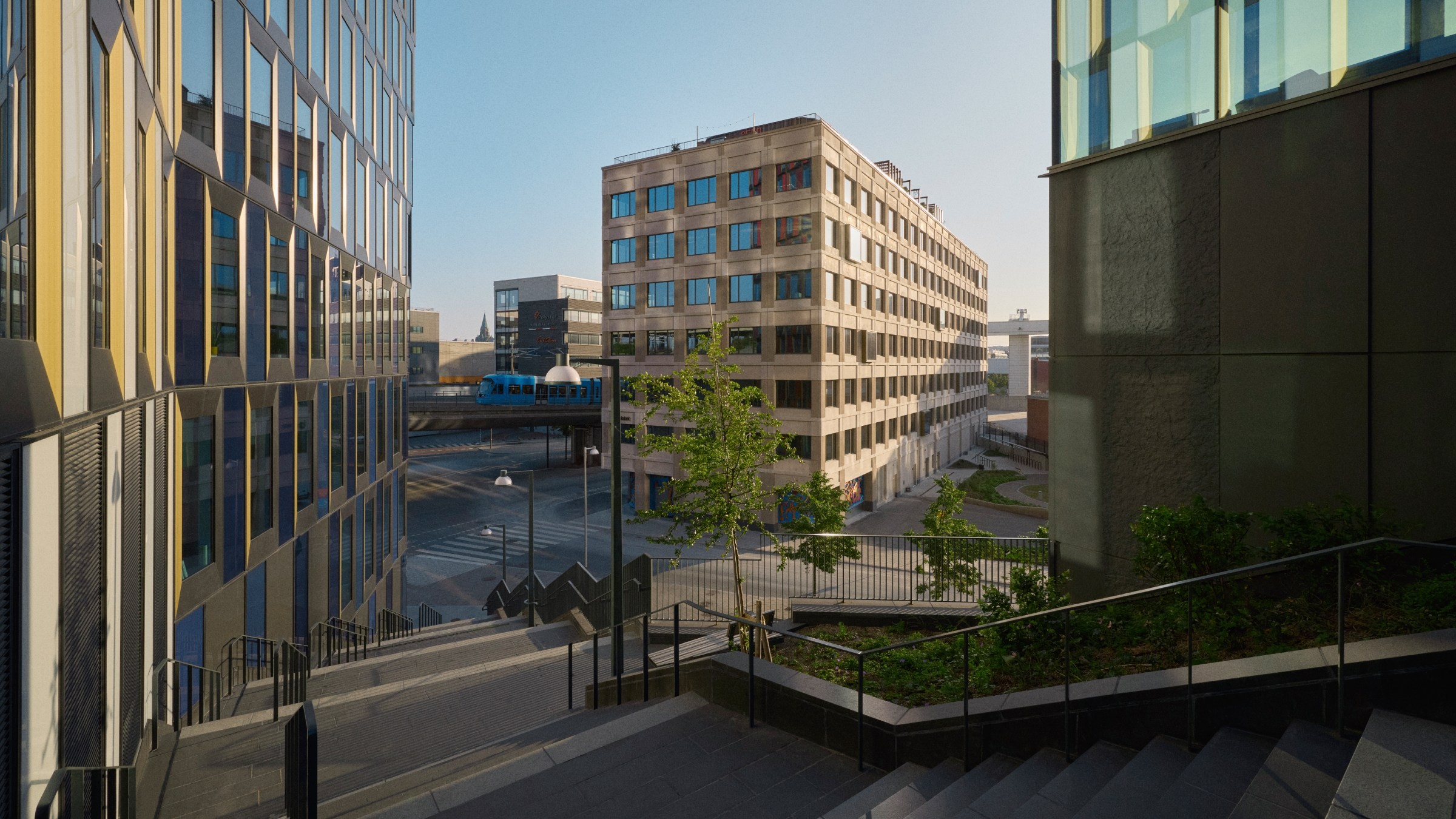 Modern stadsbild med ett tåg som passerar mellan samtida byggnader och en trappa i solsken.
