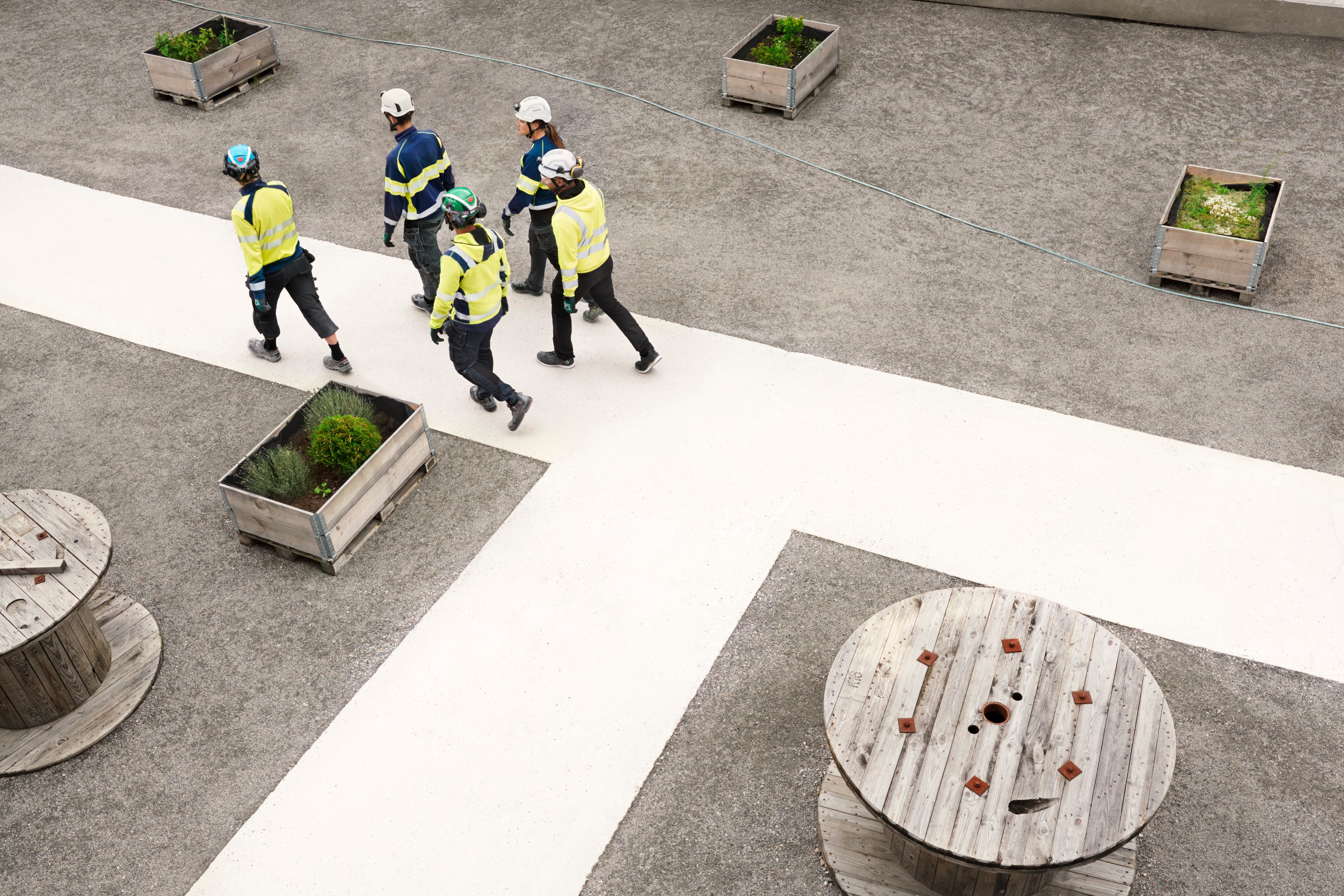 Group of workers in safety equipment walking on a concrete path with wooden pots and reels nearby.