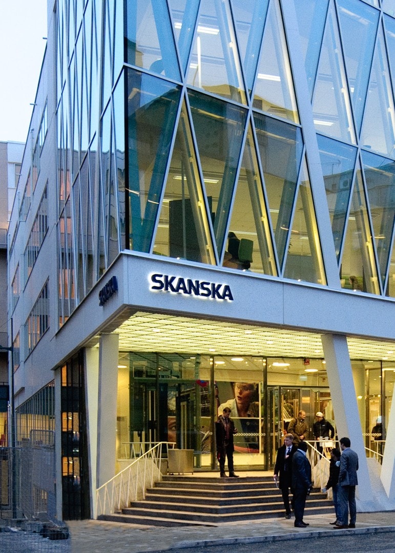Modern Skanska kontorsbyggnad med glasfasad och entré i gatunivå, kvällsvy.