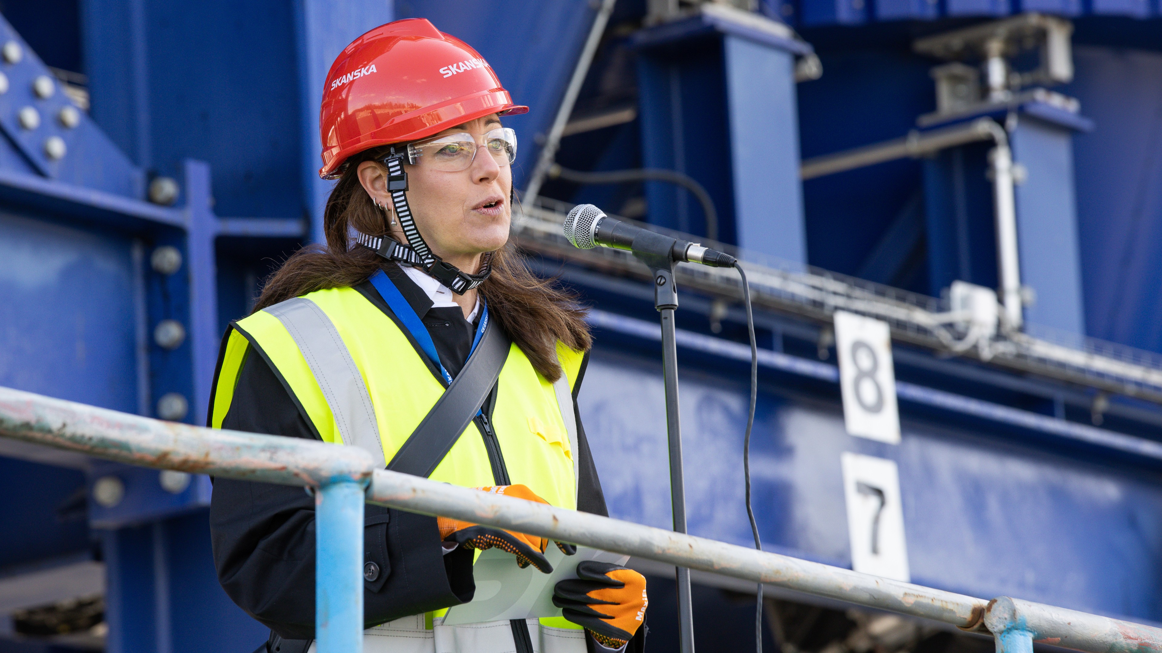 Sara Gorton hållbarhetschef Skanska. Kvinna i säkerhetsutrustning som talar i en mikrofon på en industriplats med blå strukturer.