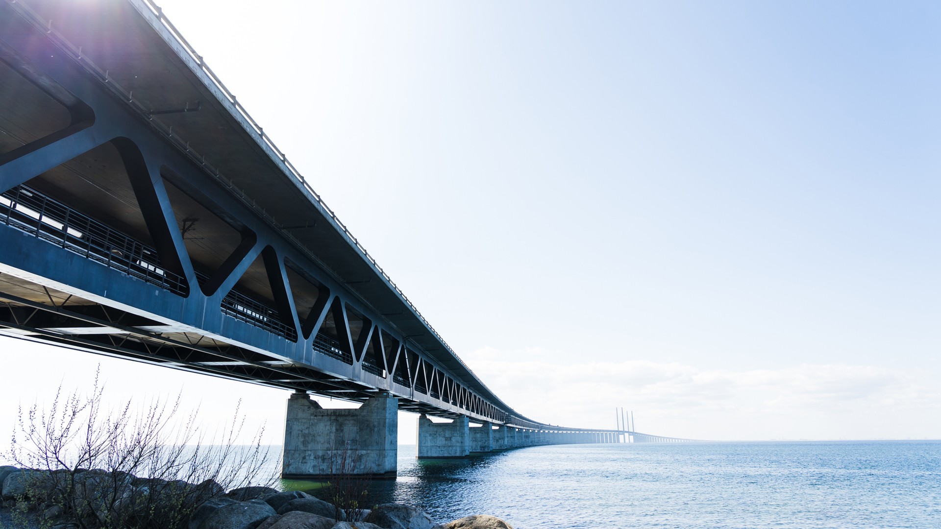 Öresundsbron har gjort det enklare att bo på ena sidan och arbeta på andra sidan. I dag arbetspendlar cirka 22 000 personer över sundet. Lång stålbro som sträcker sig över lugnt vatten under en klar blå himmel.