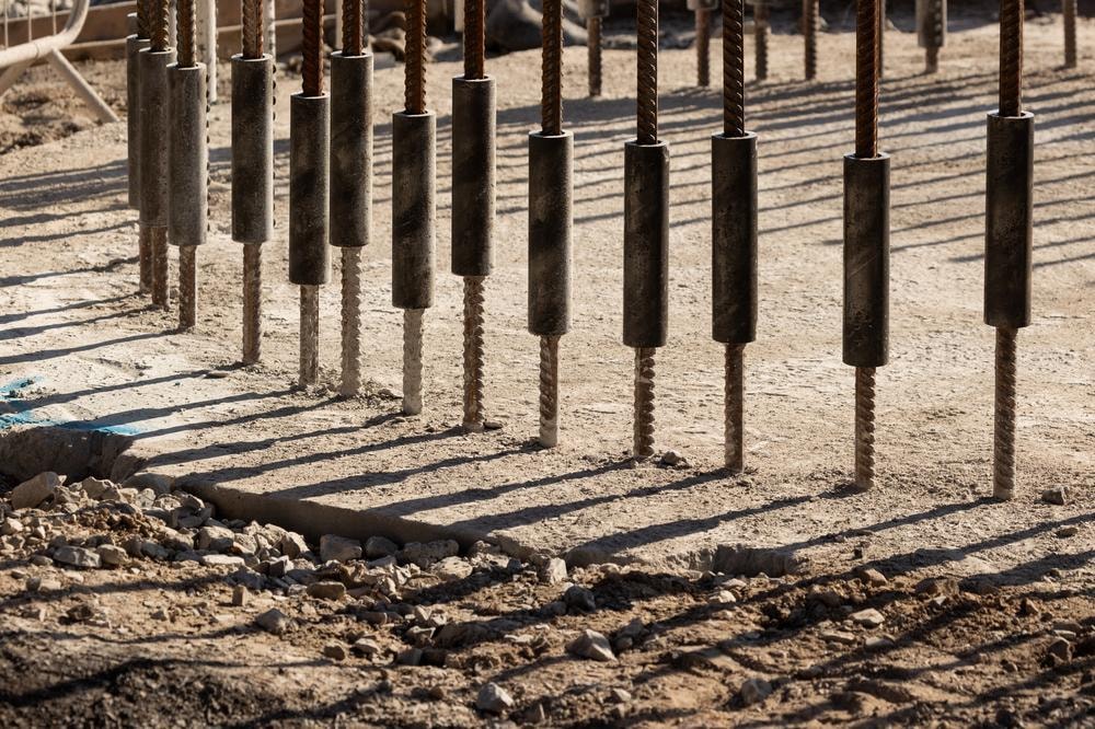 Betonowy fundament z pionowymi prętami zbrojeniowymi przygotowany do budowy, rzucający cienie na ziemię.