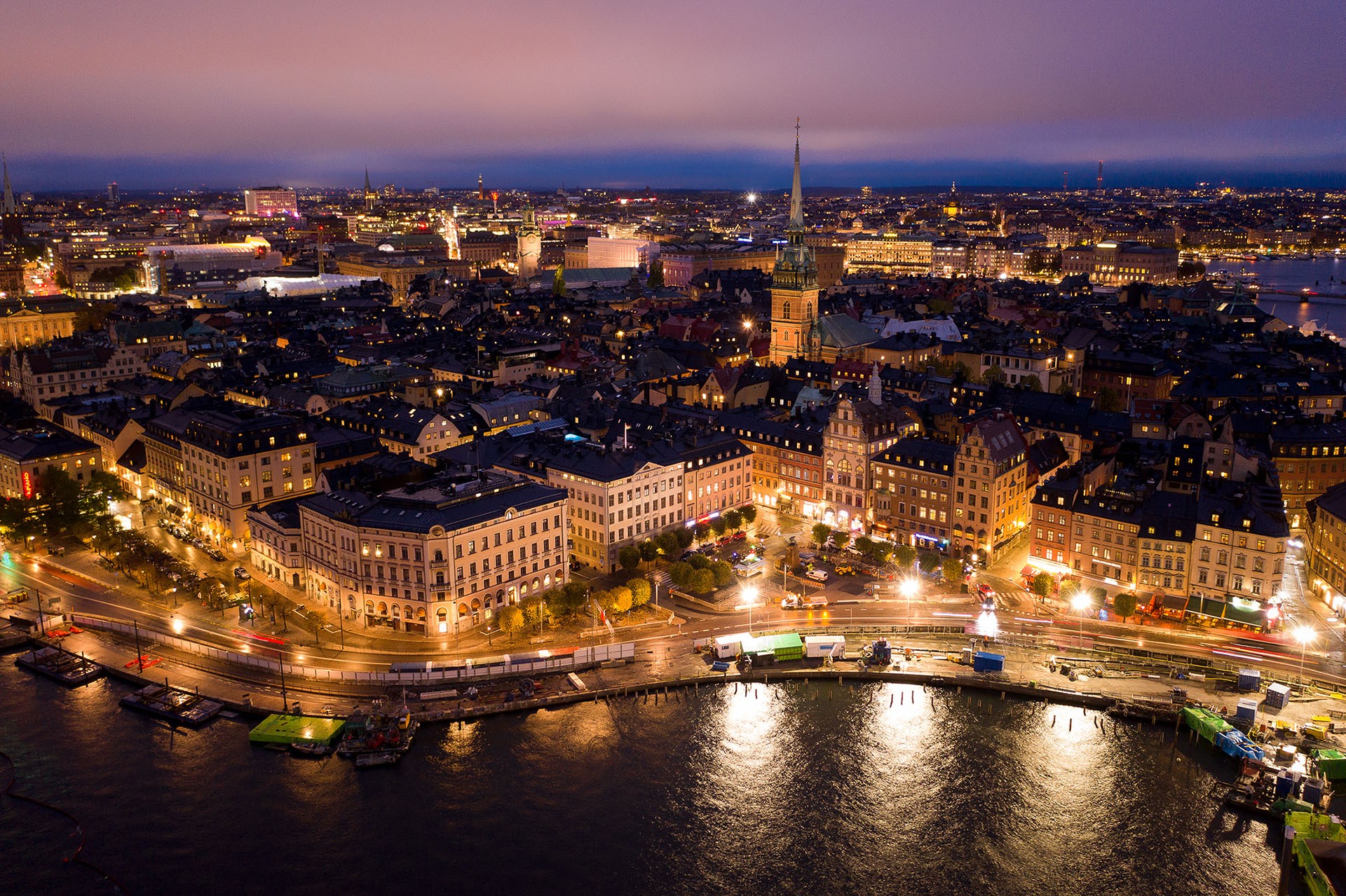 Flygfoto över Stockholm i skymningen med upplysta gator, historiska byggnader och vattenfront.