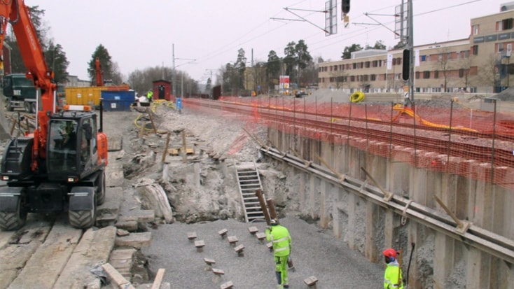 Byggeplats med arbetare och en grävmaskin nära järnvägsspår, omgiven av säkerhetsbarriärer.