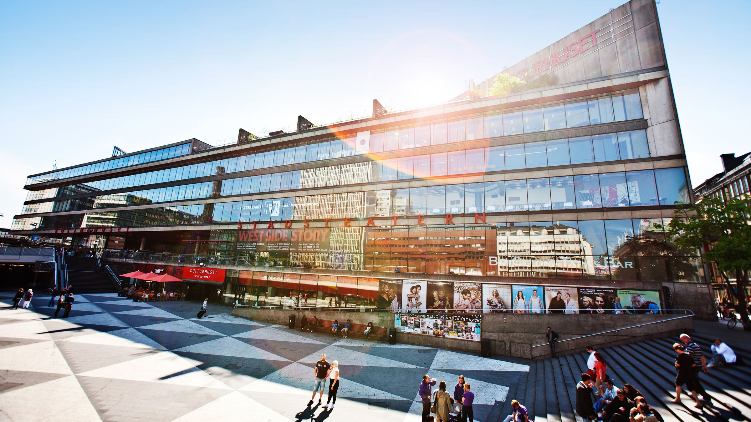 Modern glasfasad byggnad med livfulla affischer, livlig torg och solbelyst stadsmiljö.
