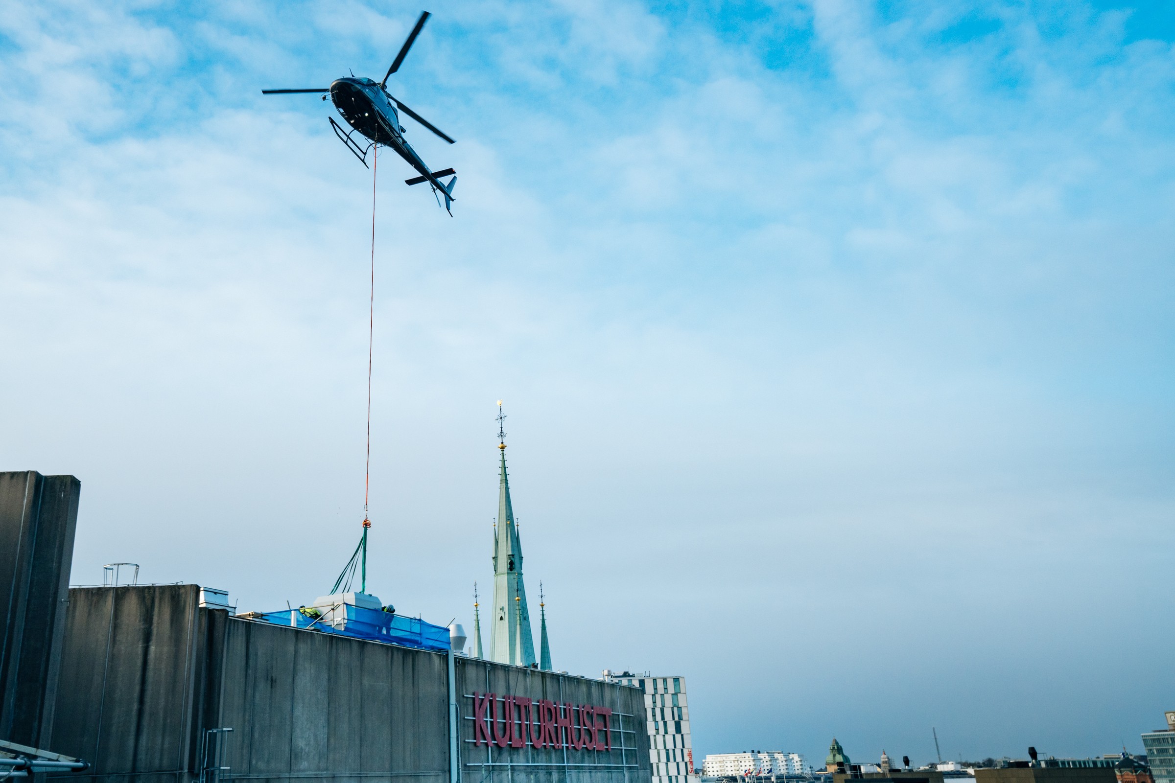 Helikopter lyfter utrustning på tak nära byggnad märkt "Kulturhuset" med kyrktorn i närheten.