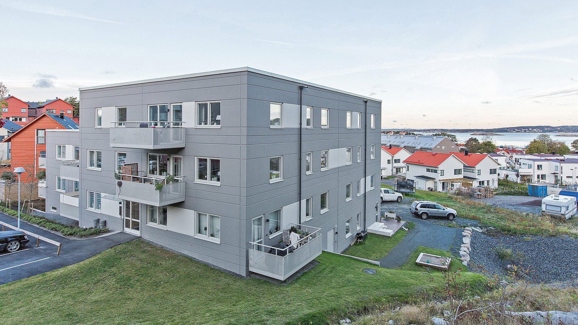 Modern lägenhetsbyggnad med balkonger, omgiven av grönska, med utsikt över en naturskön vattenfront.