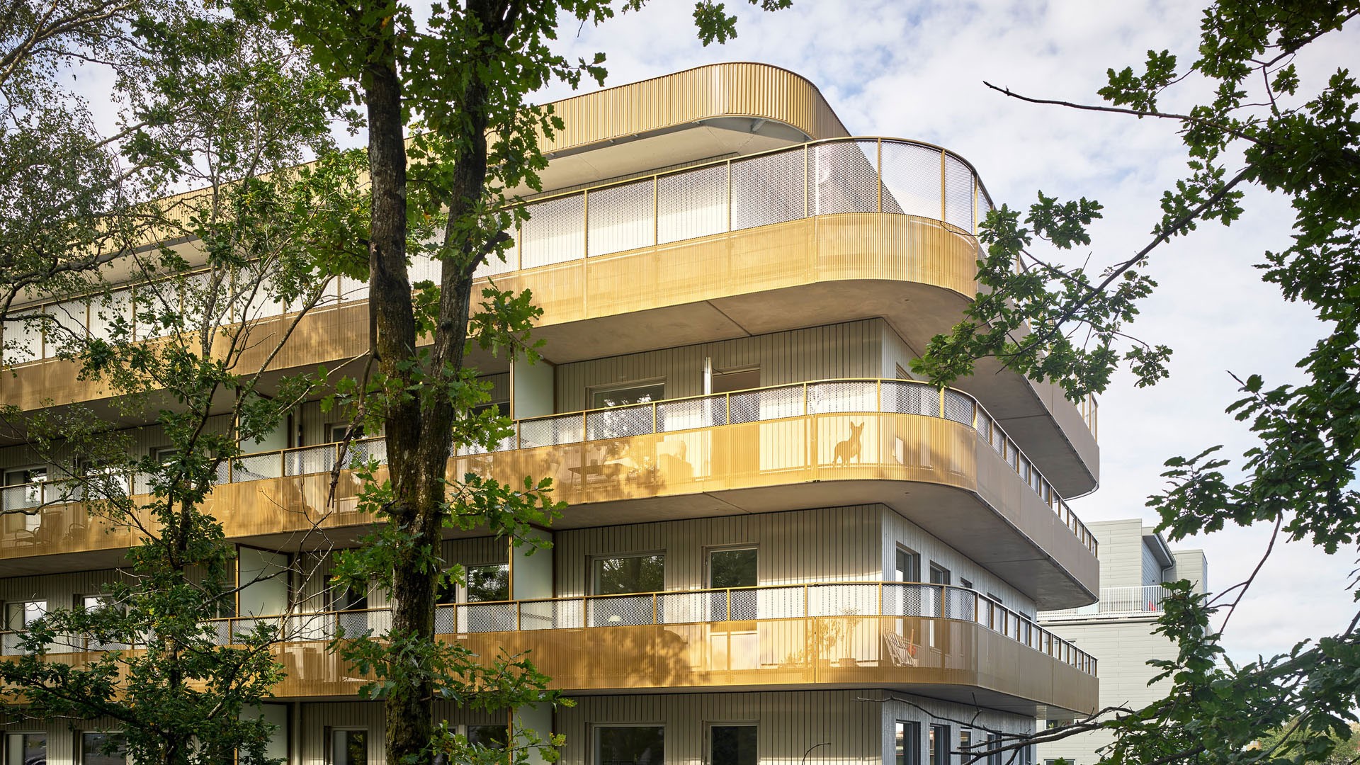 Tio år av planerad utveckling ska resultera i en stadsdel med totalt 1300 bostäder. Modern flervåningsbyggnad med böjda balkonger, omgiven av frodiga gröna träd under en molnig himmel.