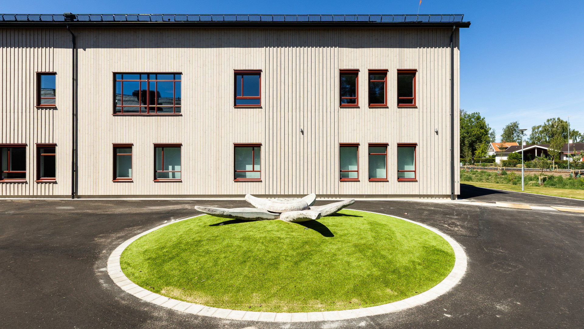 Exteriör av Västerängsskolan i Nybro med träfasad i ljus furu och röda fönster, samt konstverk på skolgården.