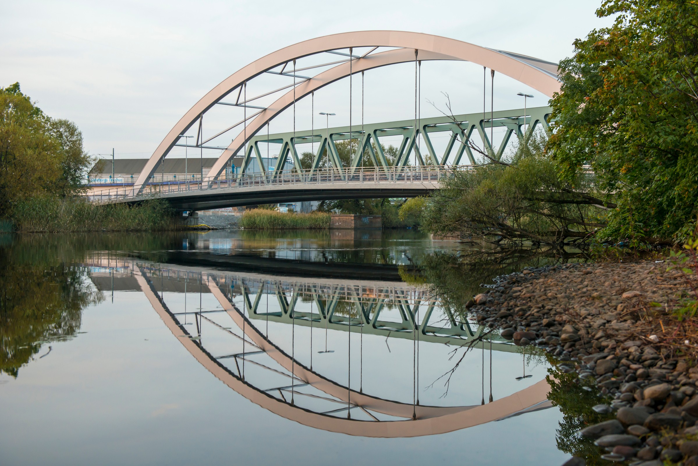 Valvbro med geometrisk design som reflekteras i lugn flod, omgiven av träd och stenar.