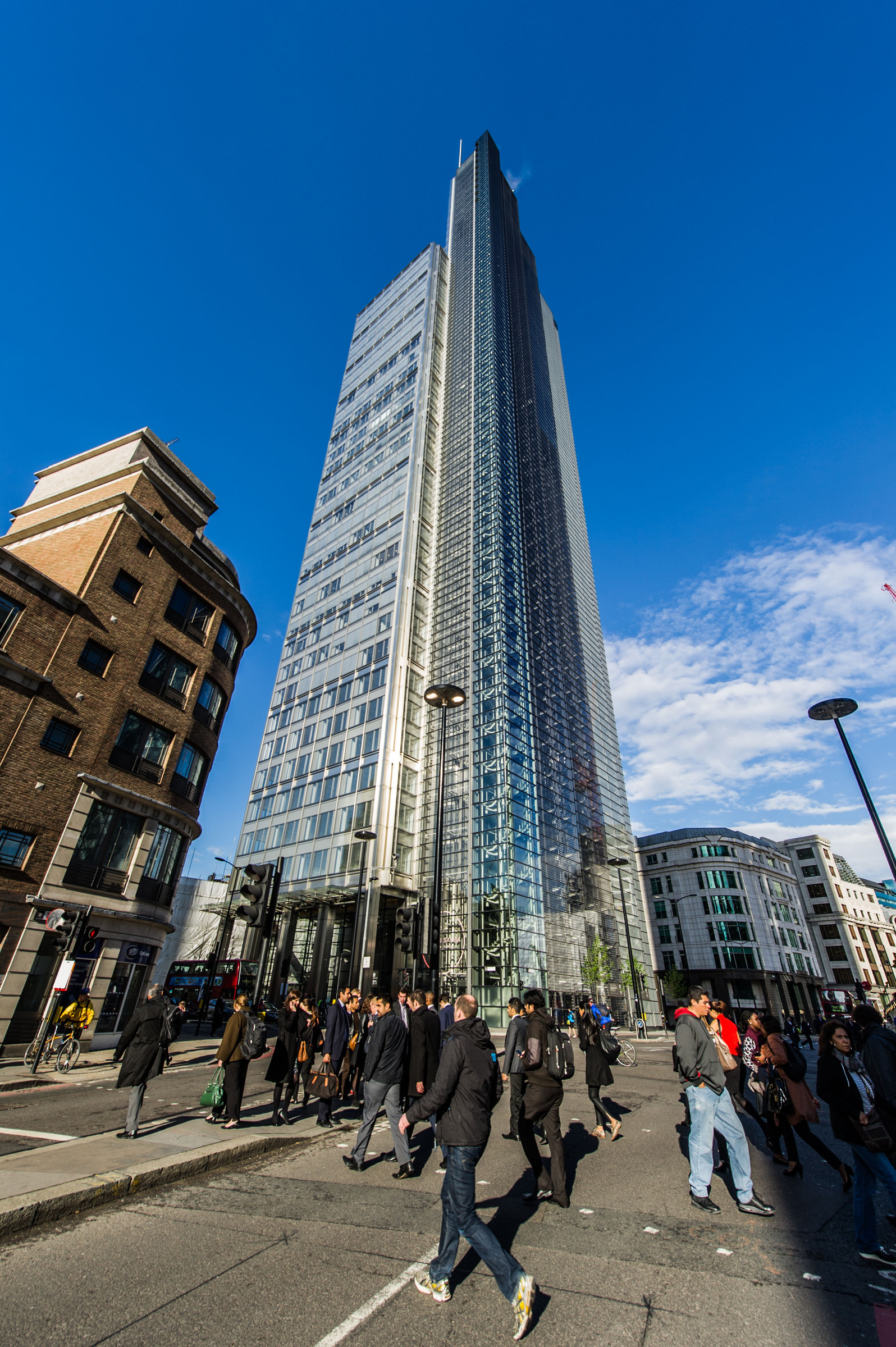 Heron Tower Skyscraper with glass facade against blue sky, surrounded by pedestrians in urban setting.