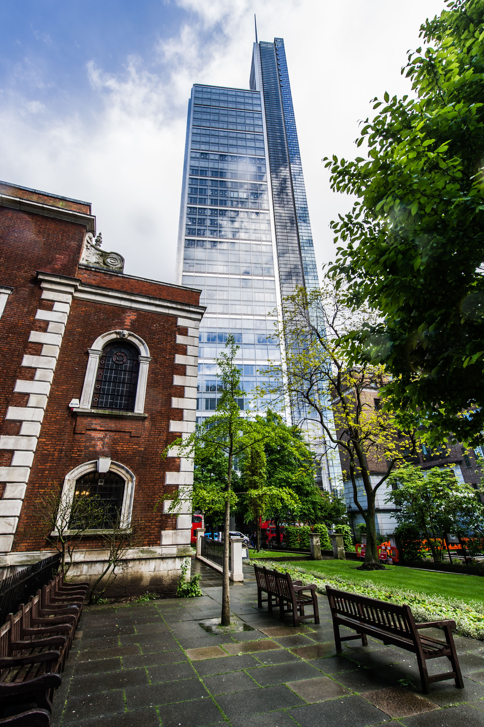 Heron Tower Modern skyscraper towers over a historic brick building and lush greenery in a cityscape.