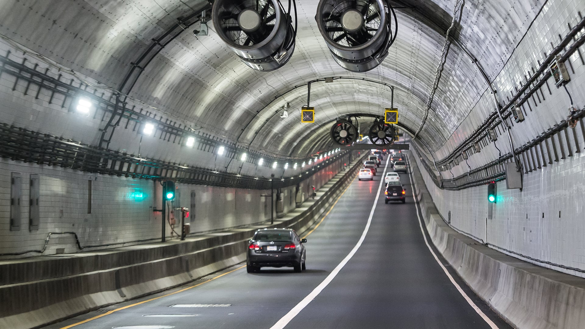 Cars Driving Through Well-Lit Urban Tunnel with Ventilation Fans Cars driving through a well-lit tunnel with ventilation fans and traffic signals.