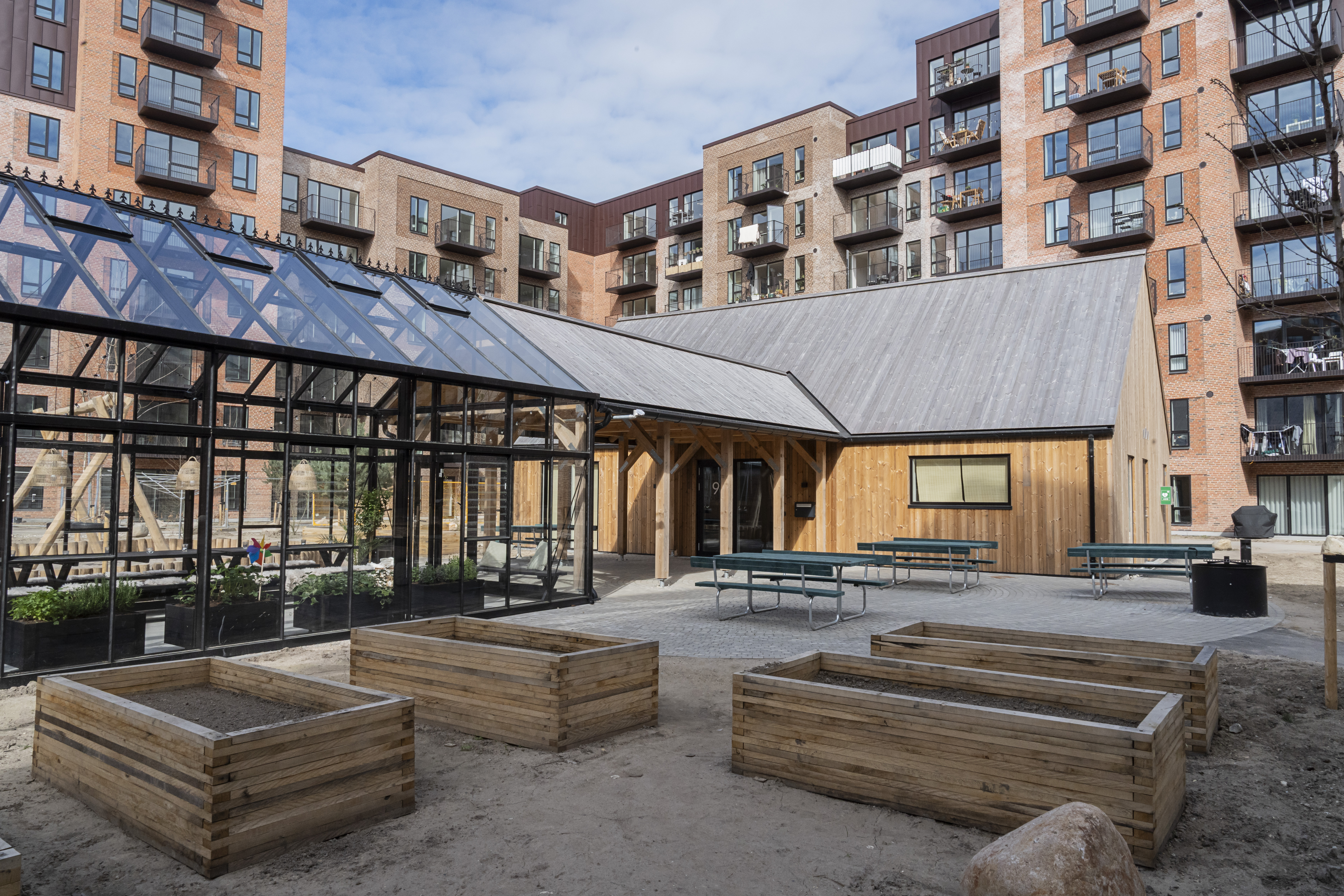 Fælleshuset i Hørkær Have Urban community garden with greenhouse and raised beds surrounded by modern apartment buildings.