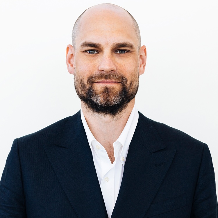 A man in a dark suit and white shirt stands against a plain white background.