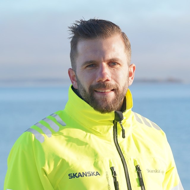 Simon Wedeborg En person i en ljusgul Skanska-jacka står utomhus med en fridfull blå havsbakgrund.