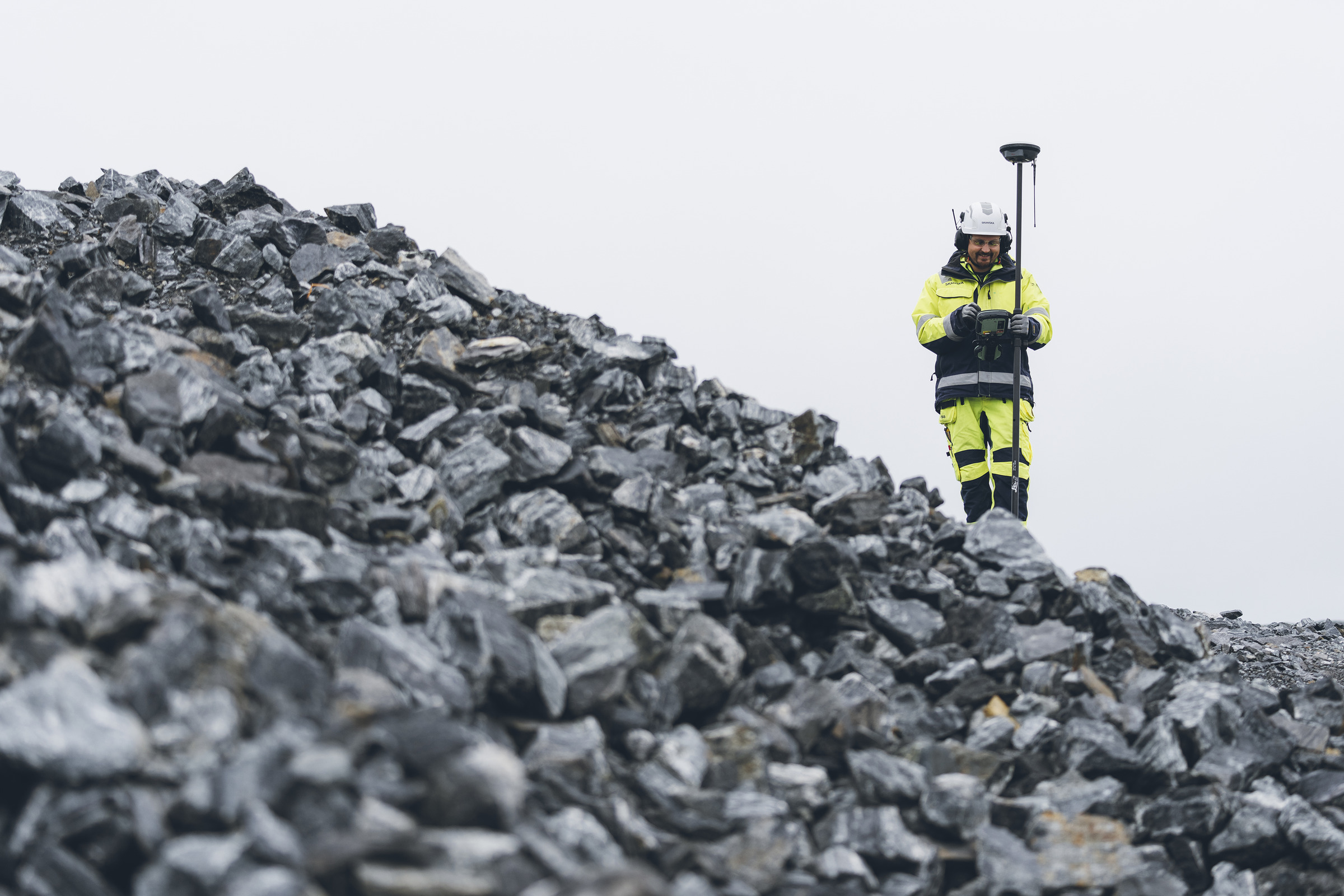 Anläggningsarbetet genomförs med en smart och hållbar massahantering för att få bra ekonomi och ekologi. Surveyor i högsynlighetsutrustning mäter terräng med GPS-utrustning på stenig landskap.